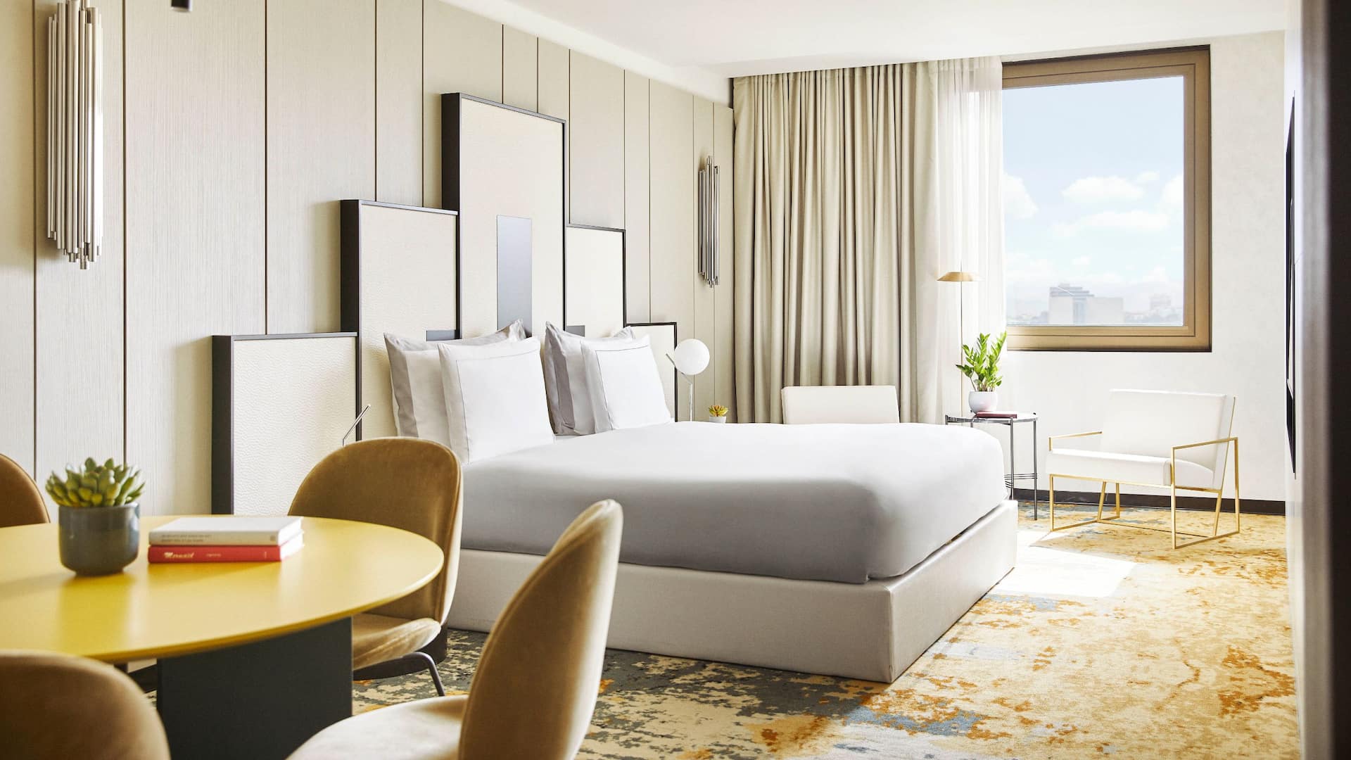 luxury hotel Studio Suite, Bed, white chairs, a round yellow table, and a carpeted floor, featuring ample natural light from a large window.
