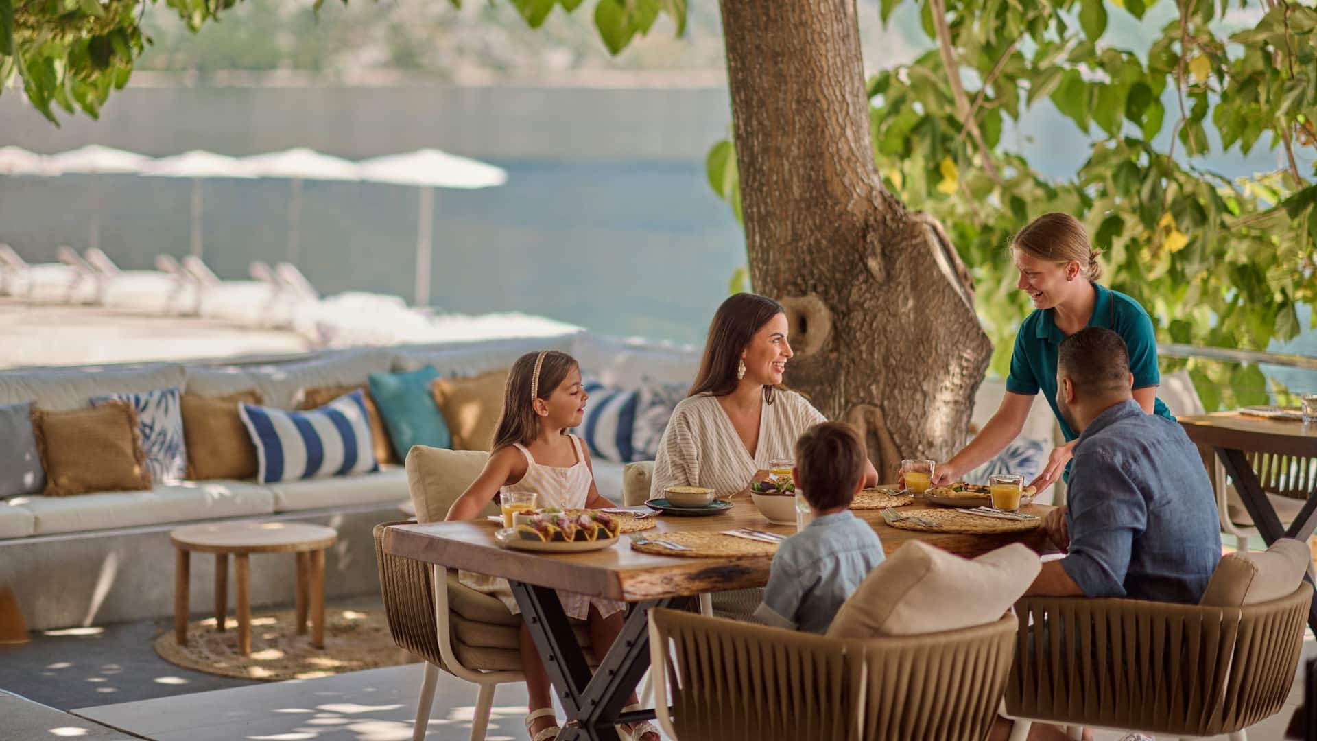 label.xOfY A family enjoying a meal together on a shaded terrace at Olé! Beach and Bar, with cozy seating, vibrant decor, and a view of the water in the background.