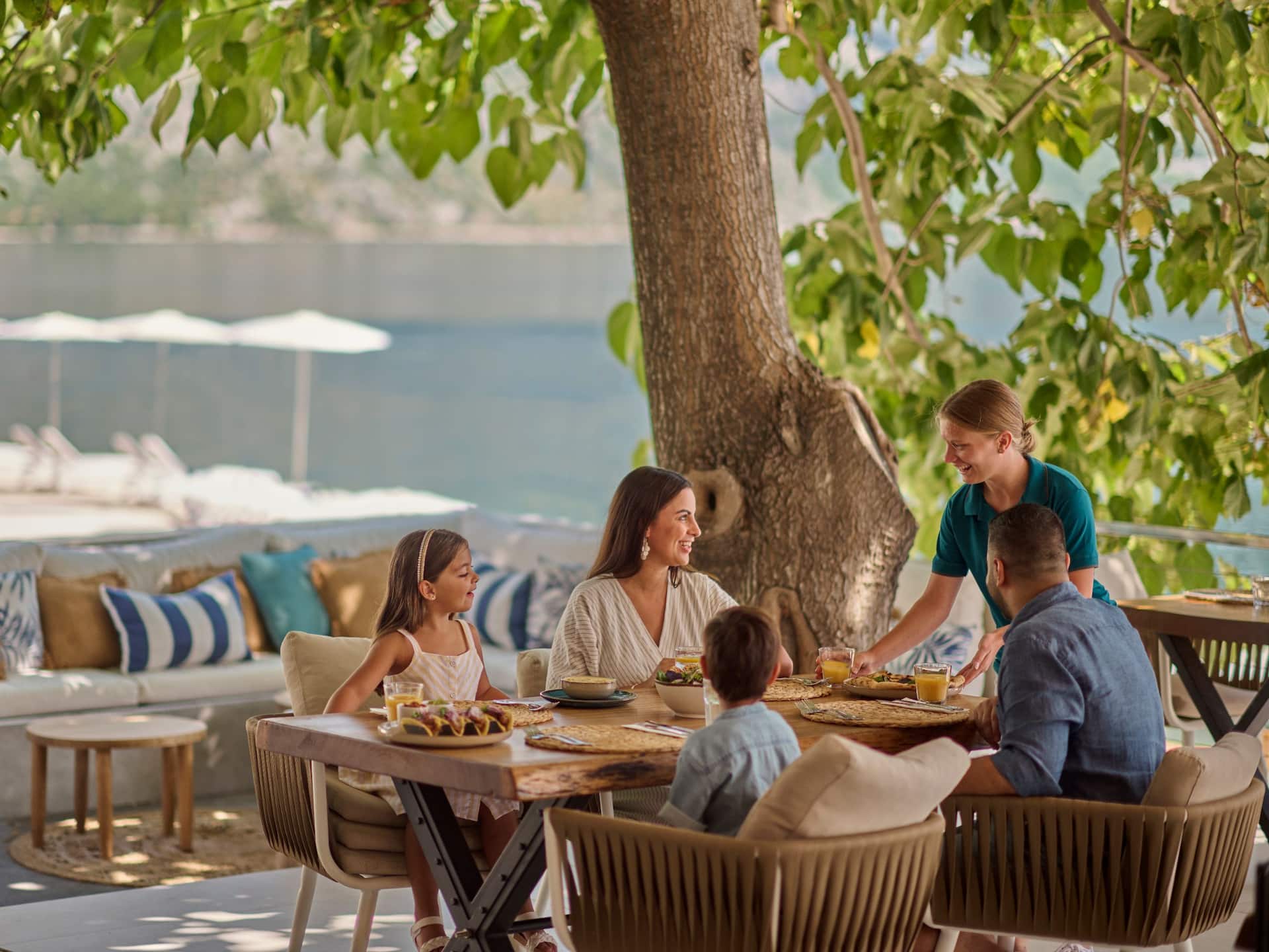 A family enjoying a meal together on a shaded terrace at Olé! Beach and Bar, with cozy seating, vibrant decor, and a view of the water in the background.