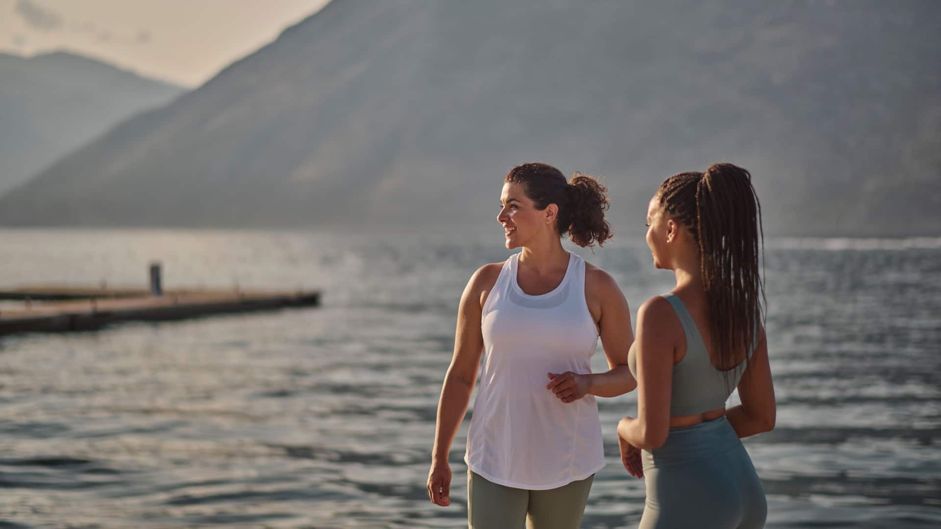Two women in activewear smiling and talking while exercising near the water at sunset, with the scenic mountains of Kotor Bay in the background.