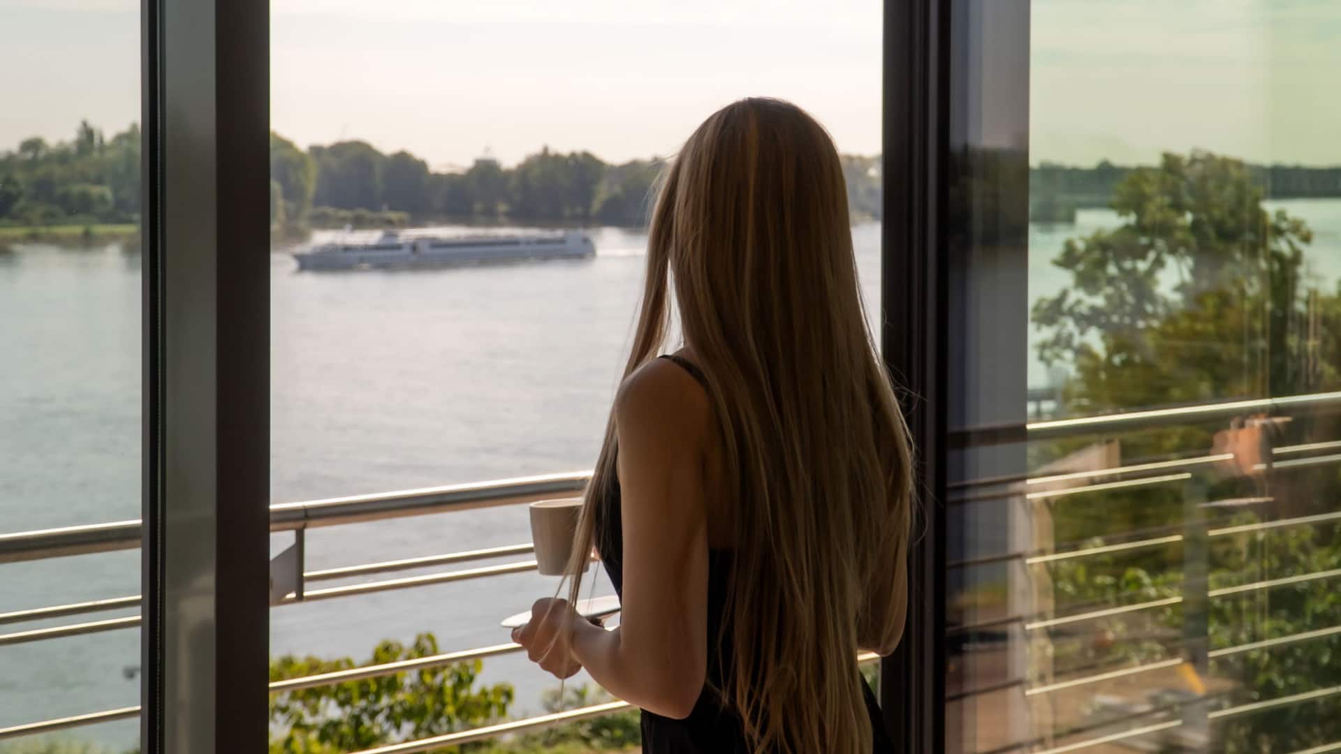 Woman in guestroom with river view standing in front of balcony watching the boats on the Rhine