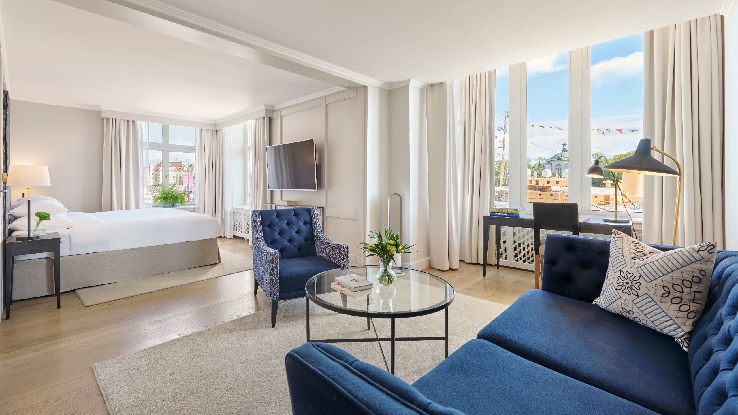 suite with a king size bed, and a seating group in blue colors on one side. Large windows with views over the water and island outside. A desk in front of one of the windows.