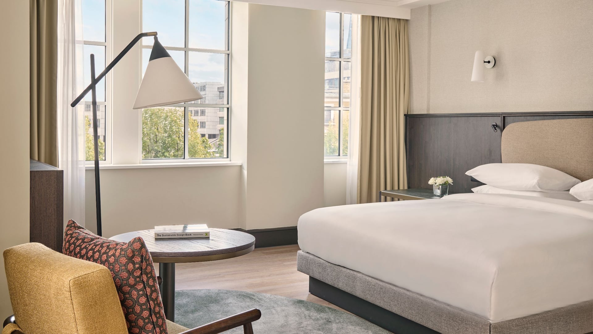 Regency Suite bedroom at Hyatt Regency London offers a king bed with white linen, a comfortable yellow chair, with an orange pillow and large windows on the City of London.