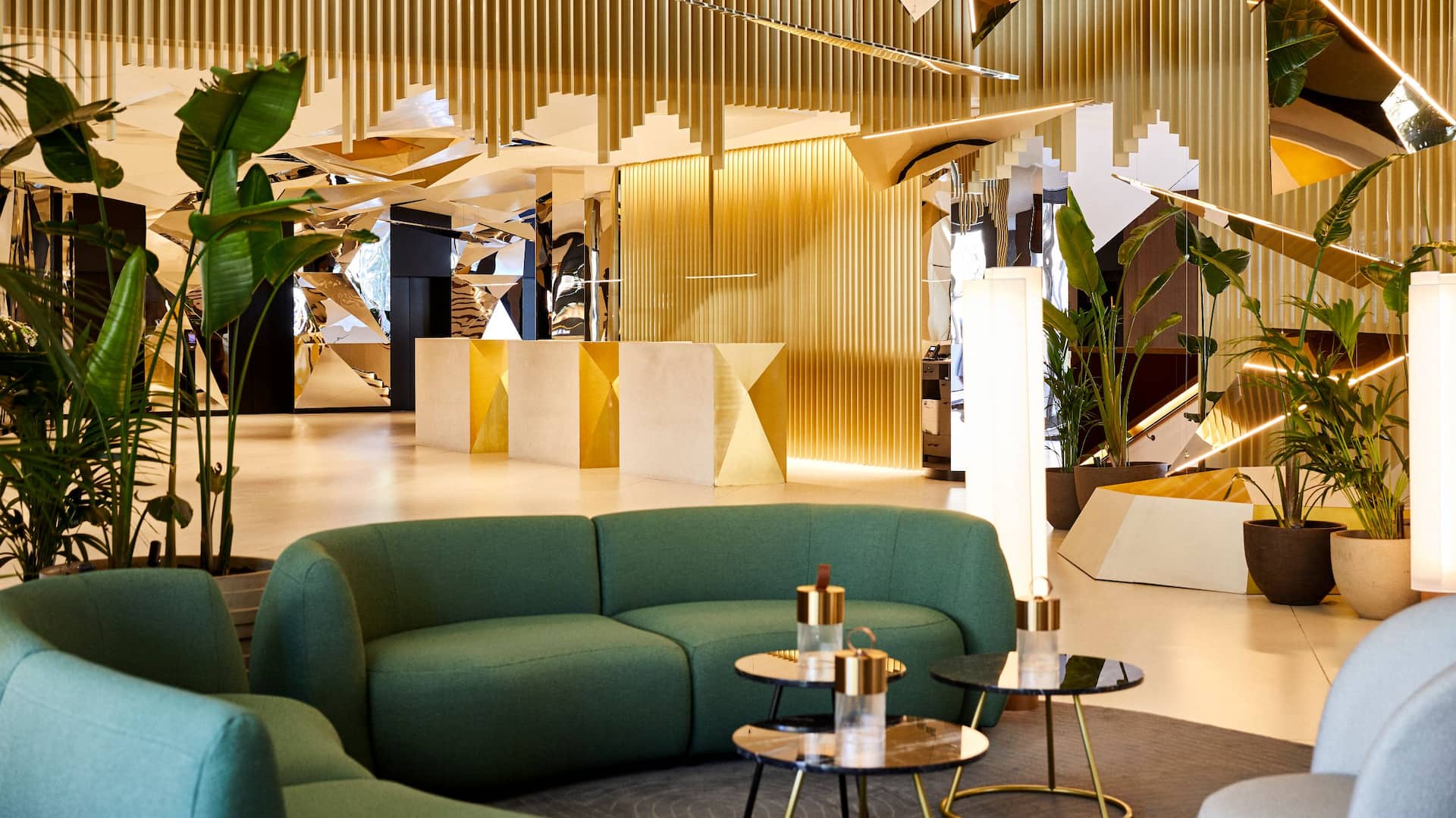 Modern hotel lobby at the Grand Hyatt Barcelona with geometric ceiling decor, green sofas, potted plants, and illuminated floor lamps arranged symmetrically. Enjoy a unique culinary experience in this five-star luxury hotel.