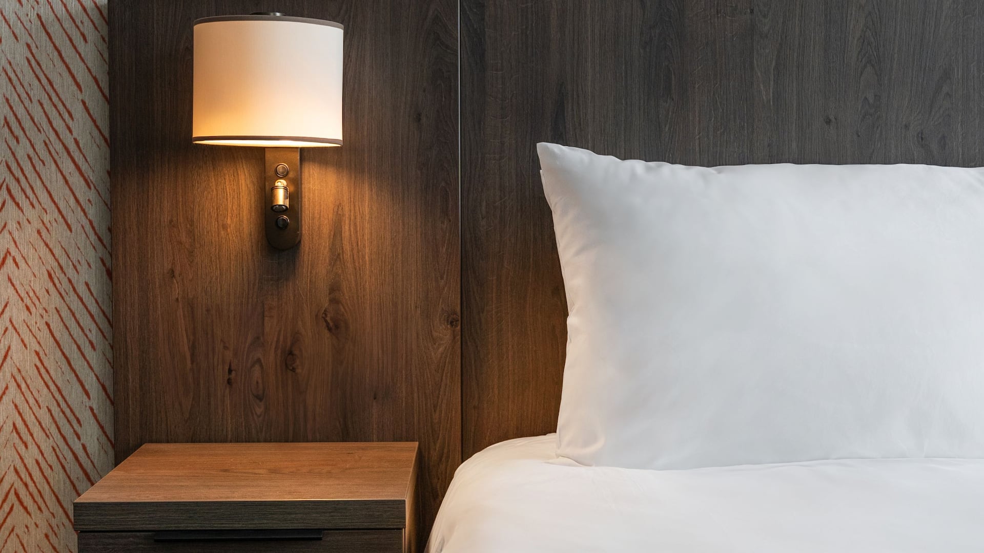 A hotel bedside table with a brass base mounted lamp and dark wood panels next to a bed with a white pillow.