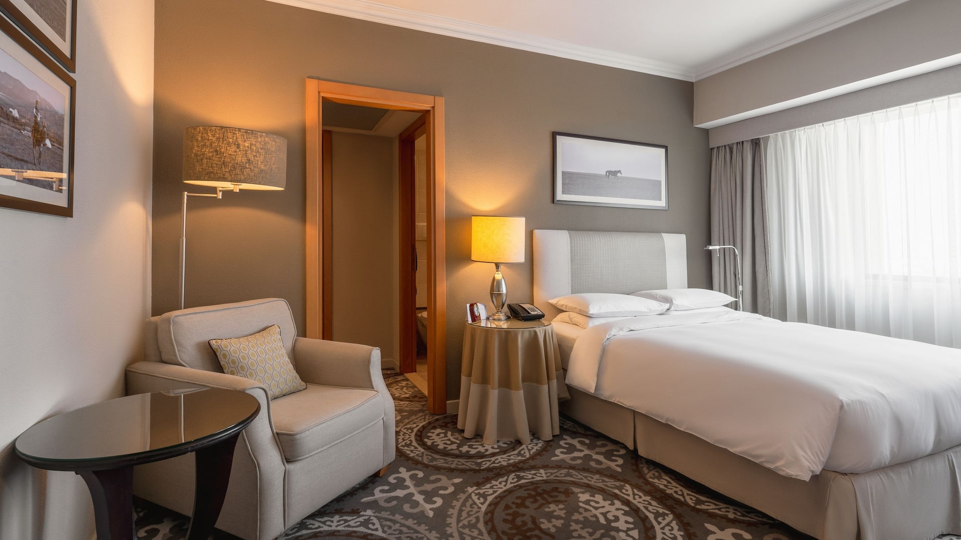 1/5 Hotel room featuring a comfortable bed, a large window with natural light, and a decorative picture on the wall, creating a welcoming and cozy ambiance