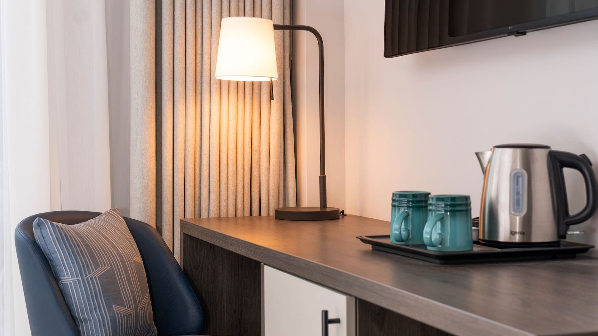 Close up of a hotel room desk with a black framed lamp, a white shade, and two teal mugs on a tray.