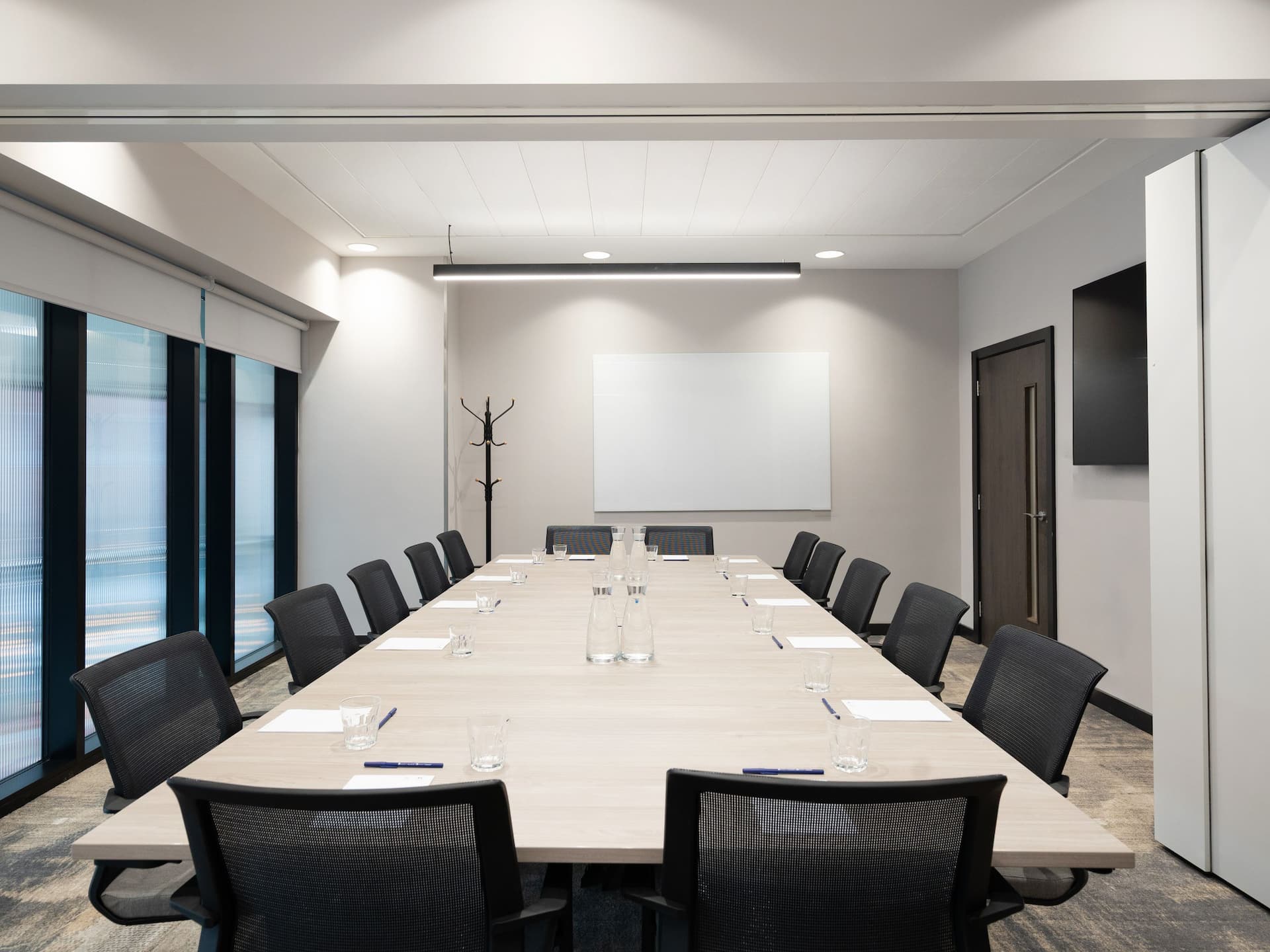 A modern, well-lit conference room with a large rectangular table, black office chairs, a whiteboard, and window blinds.
