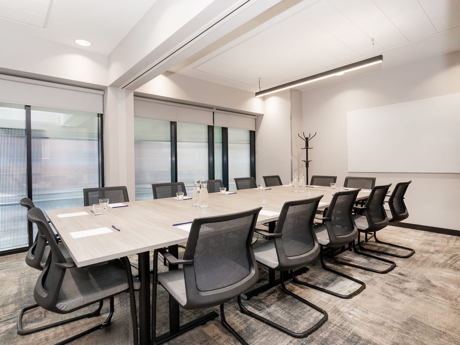 A modern conference room with a long light-coloured table surrounded by black mesh chairs featuring a whiteboard, a coat rack, and floor to ceiling windows with blinds.